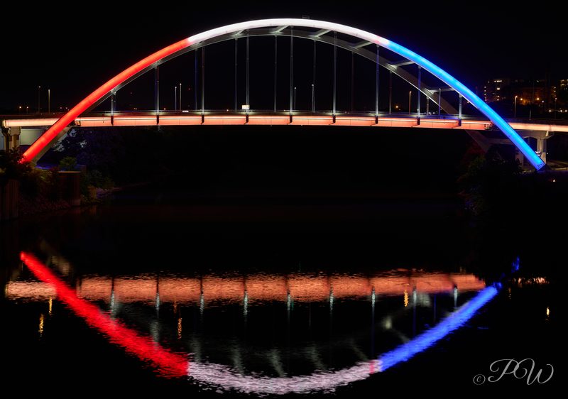 An arch bridge lit red, white and blue and reflected in the river at night