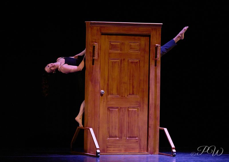 A dancer leans back beside a freestanding wooden door on a dark stage