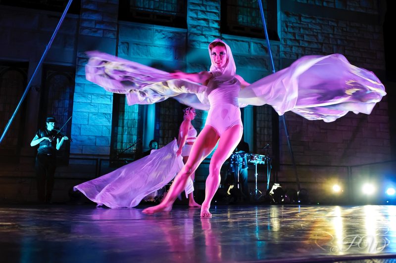 A dancer spreads pink and purple fabric wings on an outdoor stage at night