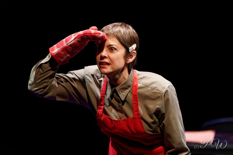 An actress in a red apron and oven mitt shades her eyes on a dark stage