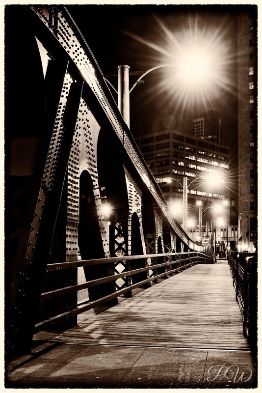A steel truss bridge walkway glowing under a streetlight at night, toned sepia