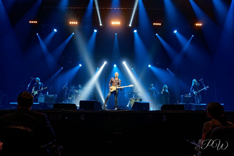 A band performing on a stage lit by blue spotlight beams, audience silhouetted in front