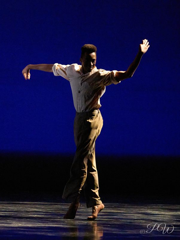 A male dancer in mid-movement with arms raised against a blue backdrop