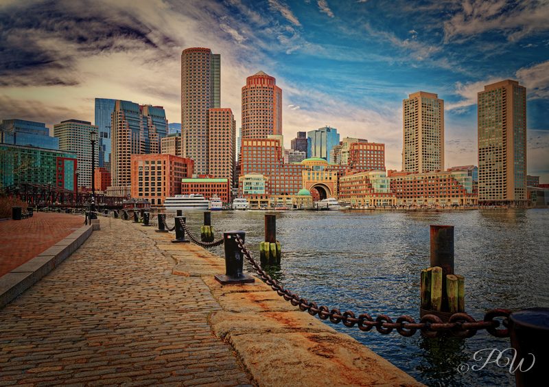 A waterfront walkway with chain posts leading toward the Boston skyline