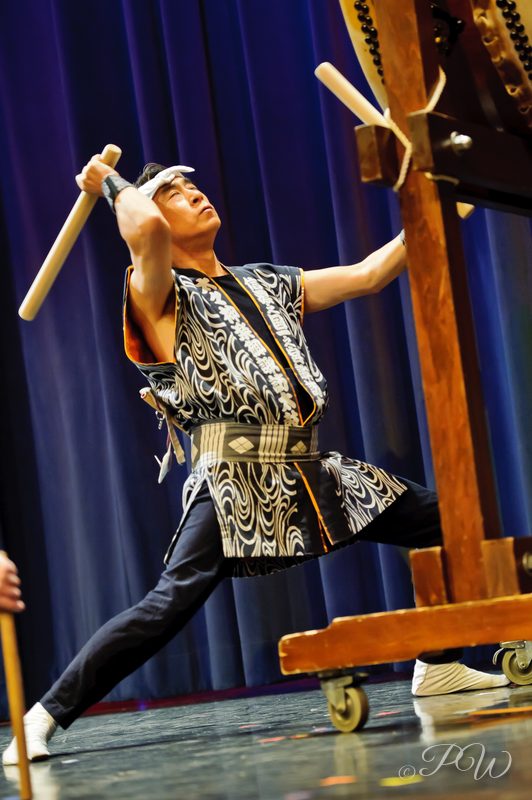 A taiko drummer in a patterned vest raises the sticks above a drum