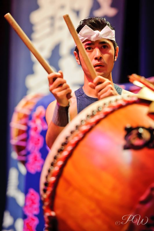 A taiko drummer in a headband plays a large barrel drum