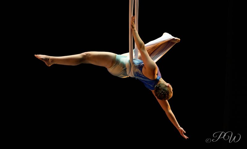 An aerialist holds a split while hanging from a trapeze against black