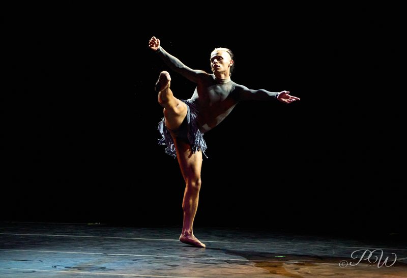 A dancer balances on one leg with the other raised high on a dark stage
