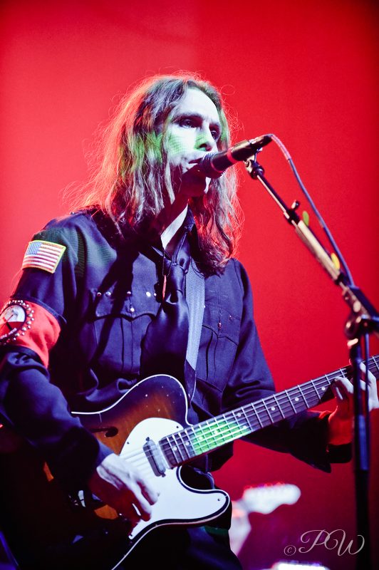 A long-haired guitarist singing into a microphone against a red backdrop