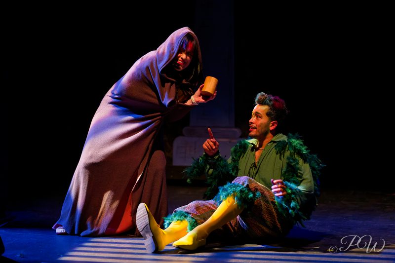 A robed figure leans over a costumed actor sitting on the stage floor