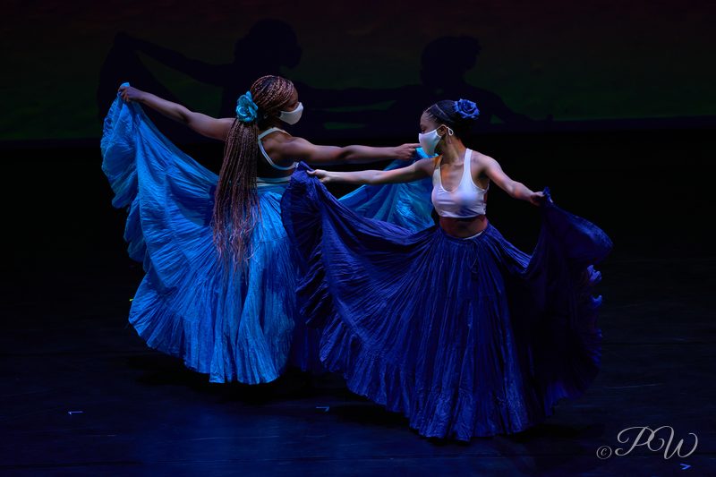 Two dancers swirl long blue skirts on a dark stage