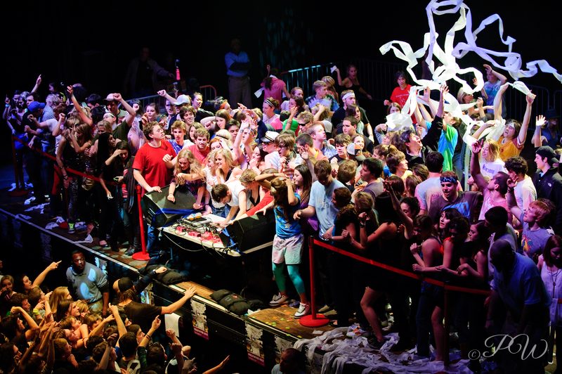 A packed crowd dancing on stage under flying streamers at a Girl Talk show