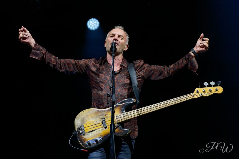 A singer with a bass guitar spreads his arms wide on a dark stage