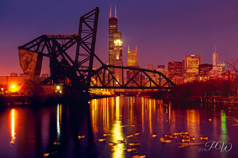 A raised railway lift bridge in front of the lit Chicago skyline and river at night