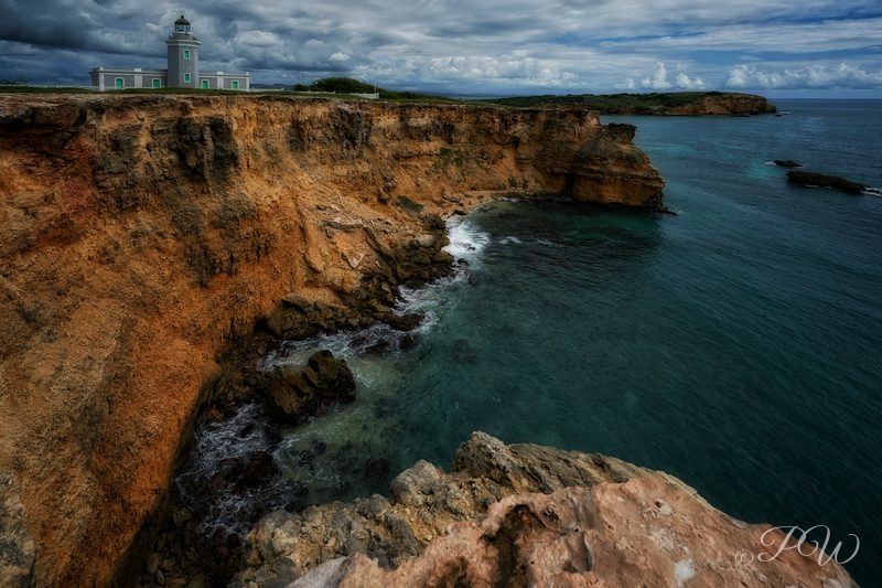 Rocky coastal cliffs above a turquoise sea with a white lighthouse on the headland