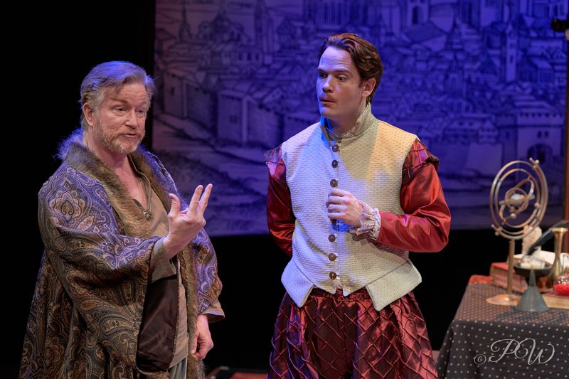 Two actors in period costume talk on a painted stage set