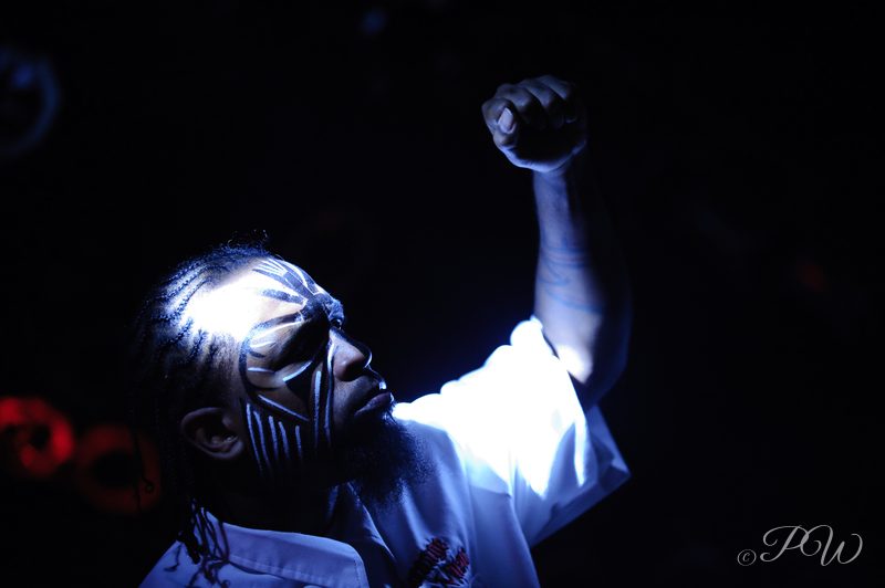 Tech N9ne in face paint raises a fist under a blue spotlight