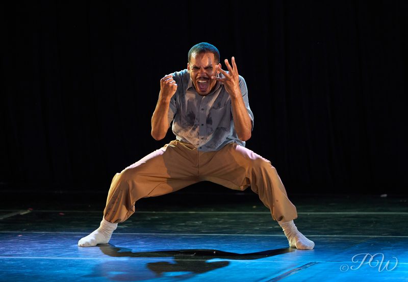 A dancer crouches low with an intense expression on a blue-lit stage