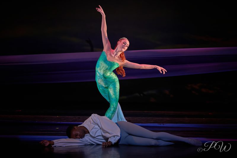 A dancer in a teal costume rises above a partner lying on the stage