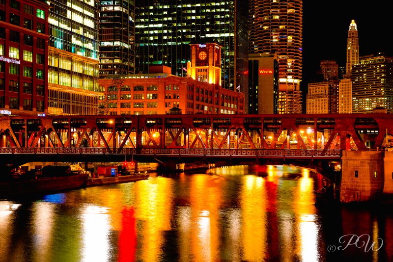 A lit elevated train bridge and towers reflected in the Chicago River at night