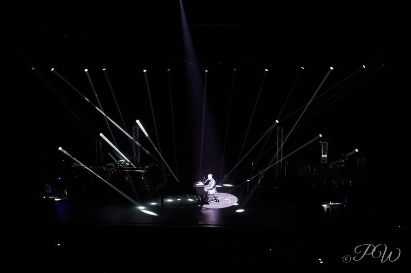 White light beams criss-cross above a lone performer on a dark stage
