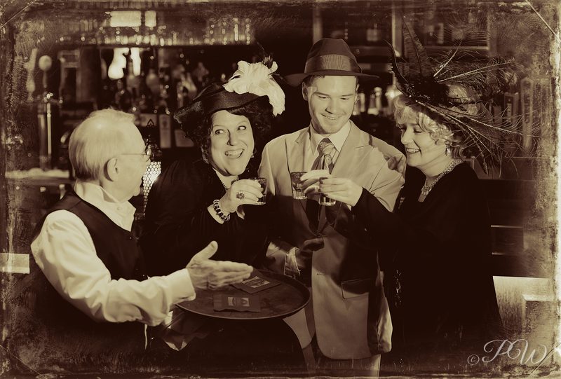 Actors in period dress gathered around a table, toned sepia like an old photograph